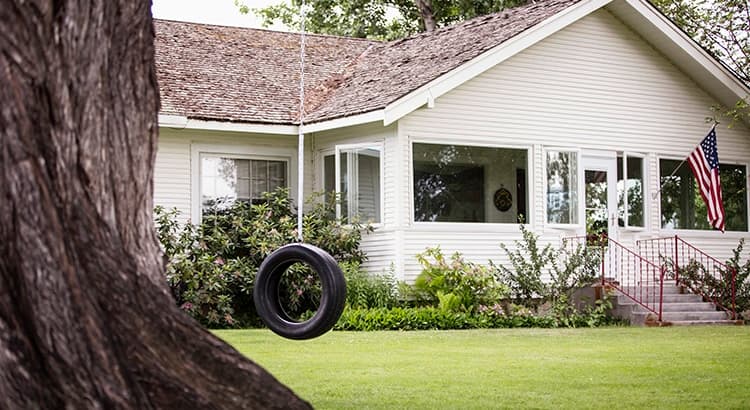 A charming white house with a shingled roof and large windows sits behind a green lawn. An American flag hangs by the front steps, while a tire swing sways from a tree in the yard. Lush shrubs and plants line the welcoming home.
