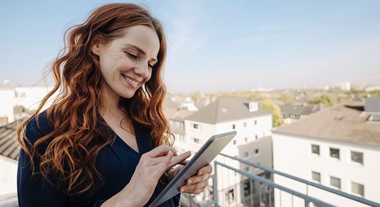A woman with long red hair smiles while using a tablet on a balcony overlooking rooftops, perhaps reading about whether the housing market will crash, on a sunny day.