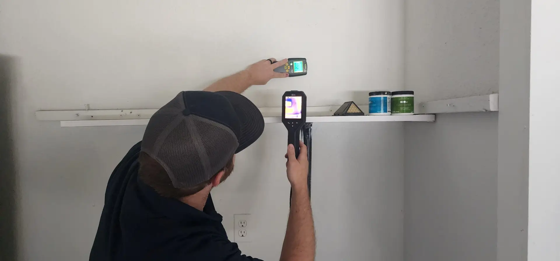A person in a dark shirt and cap uses a handheld thermal imaging device to scan a white wall near a shelf with containers. Another device is held up, its screen displaying detailed imaging results.