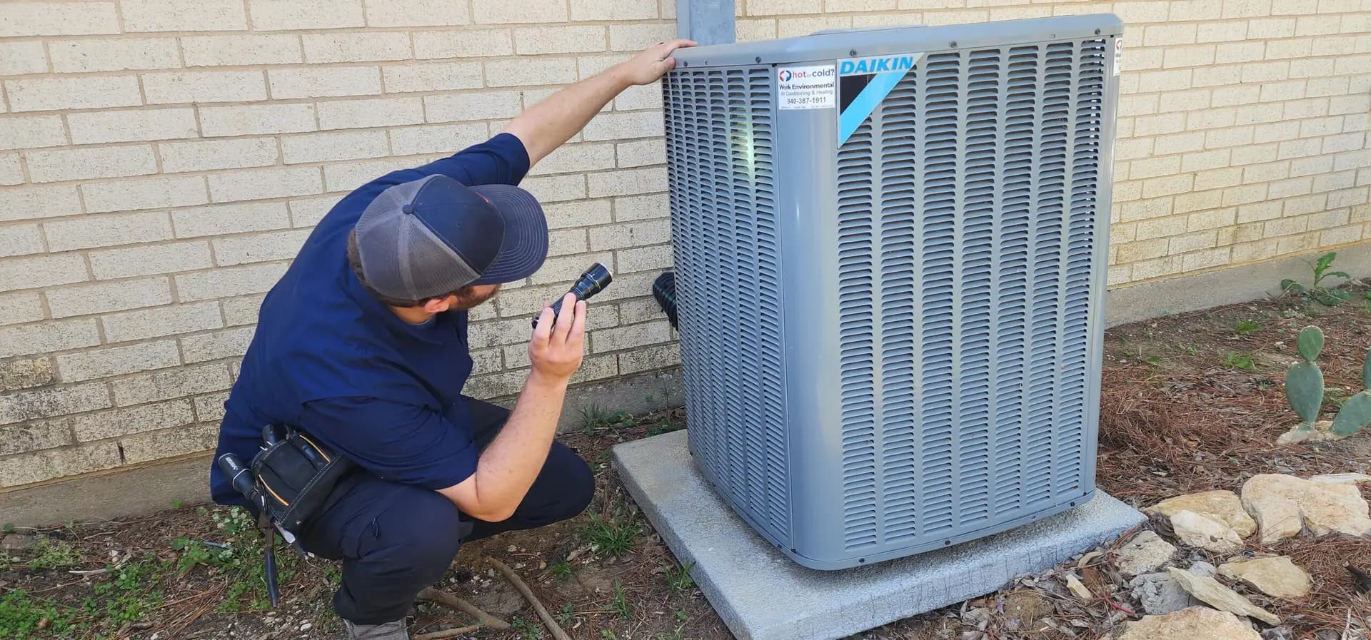 A technician in a navy uniform and cap inspects an outdoor Daikin air conditioning unit with a handheld device, crouching beside a brick wall. The focus is on the detailed maintenance work.