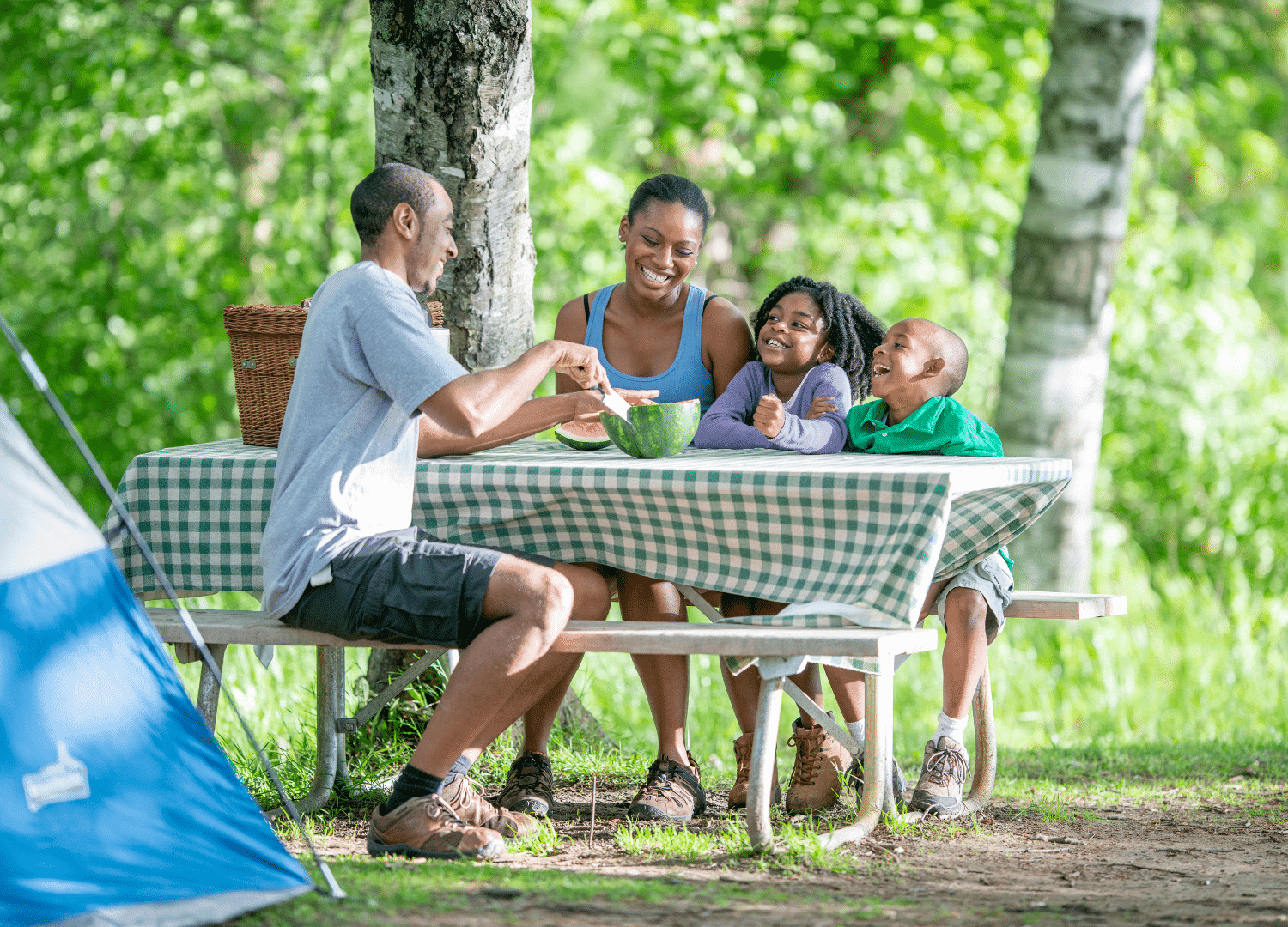 A family of four sits at a picnic table outdoors, smiling and enjoying a meal together. Trees and greenery surround them, and a tent is visible nearby, highlighting their camping adventure.