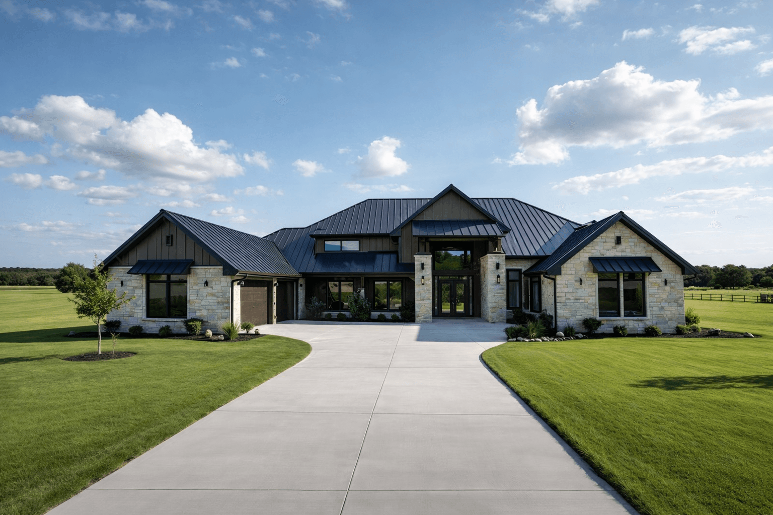 A modern ranch-style house with a dark metal roof, stone exterior, and large windows stands amid landscaped lawns, set against a vibrant green field and blue sky dotted with clouds.