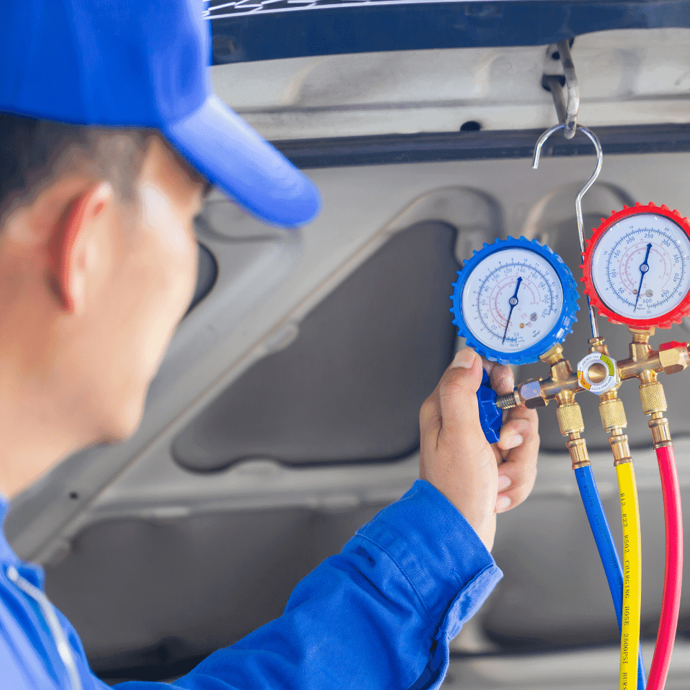 A person wearing a blue uniform is checking AC pressure with HVAC manifold gauges, using blue, red, and yellow hoses to inspect an air conditioning system under the hood of a vehicle.