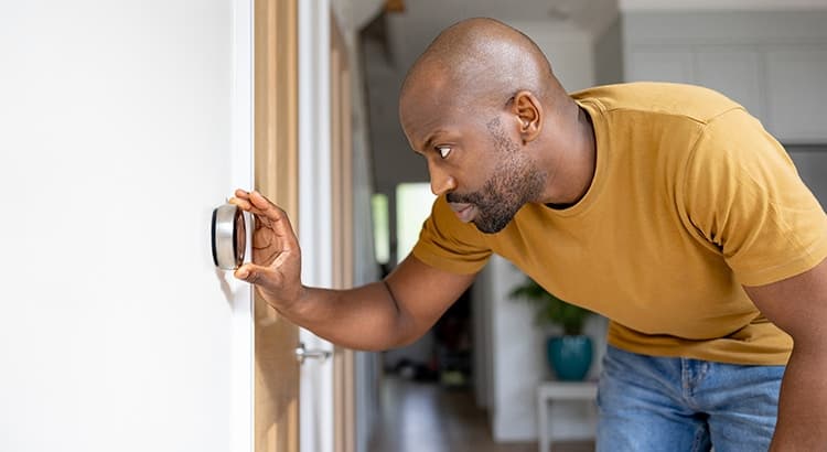 A man in a mustard yellow shirt adjusts a modern smart thermostat on a white wall inside a bright, contemporary home, possibly considering an AC replacement. He leans forward, looking closely at the device.