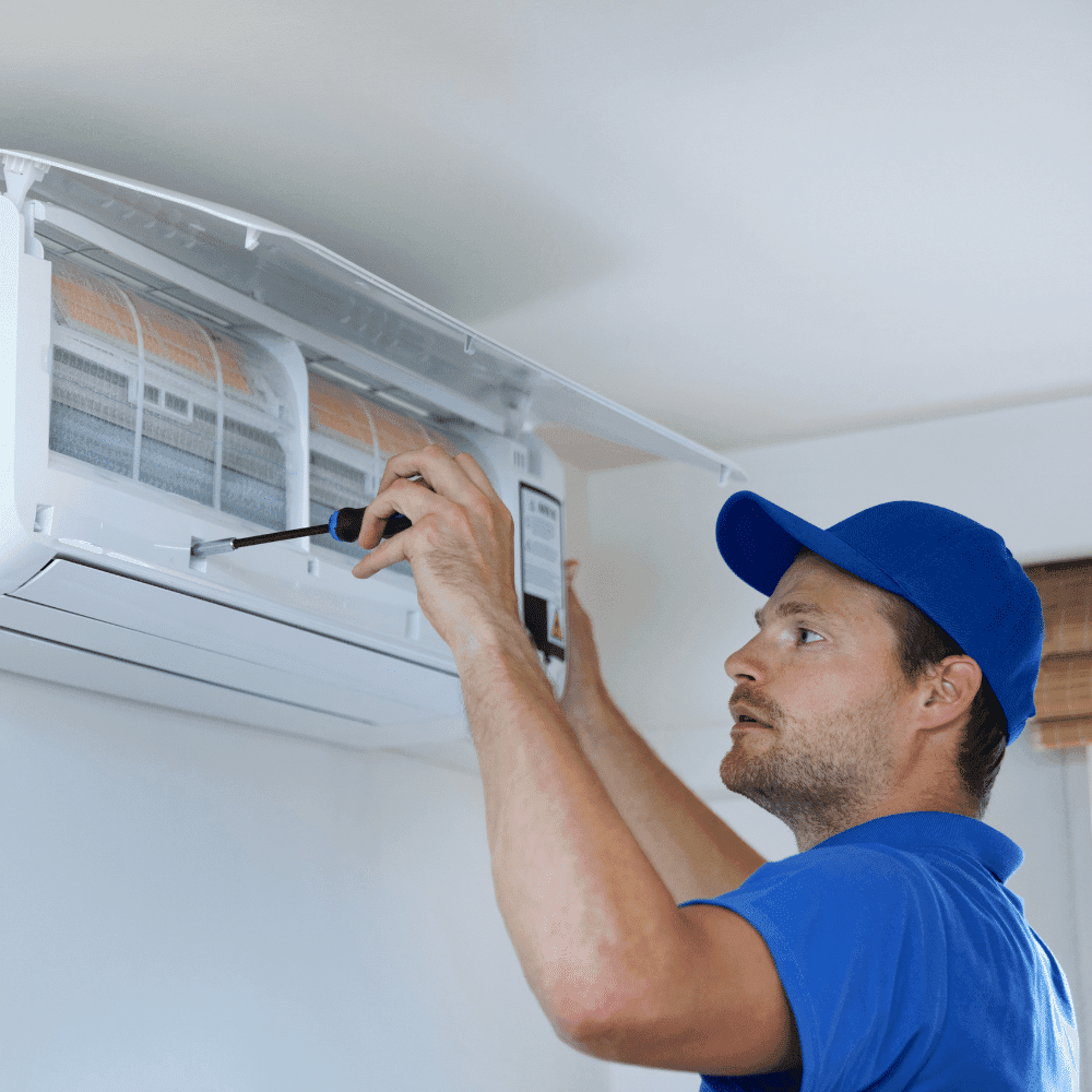 A technician in a blue shirt and cap is carefully installing or repairing a wall-mounted air conditioner unit with a screwdriver inside a room.