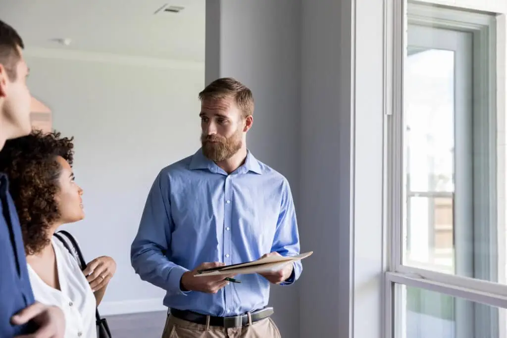 A man holding a folder stands by a window, discussing the importance of home inspection with a woman and another man inside a bright, modern room.