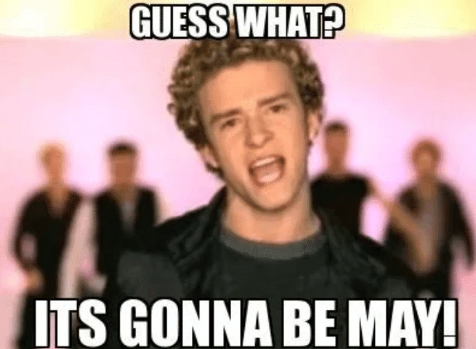 A man with curly hair sings, with bold white text above and below him reading, GUESS WHAT? ITS GONNA BE MAY! The scene features people standing behind him.
