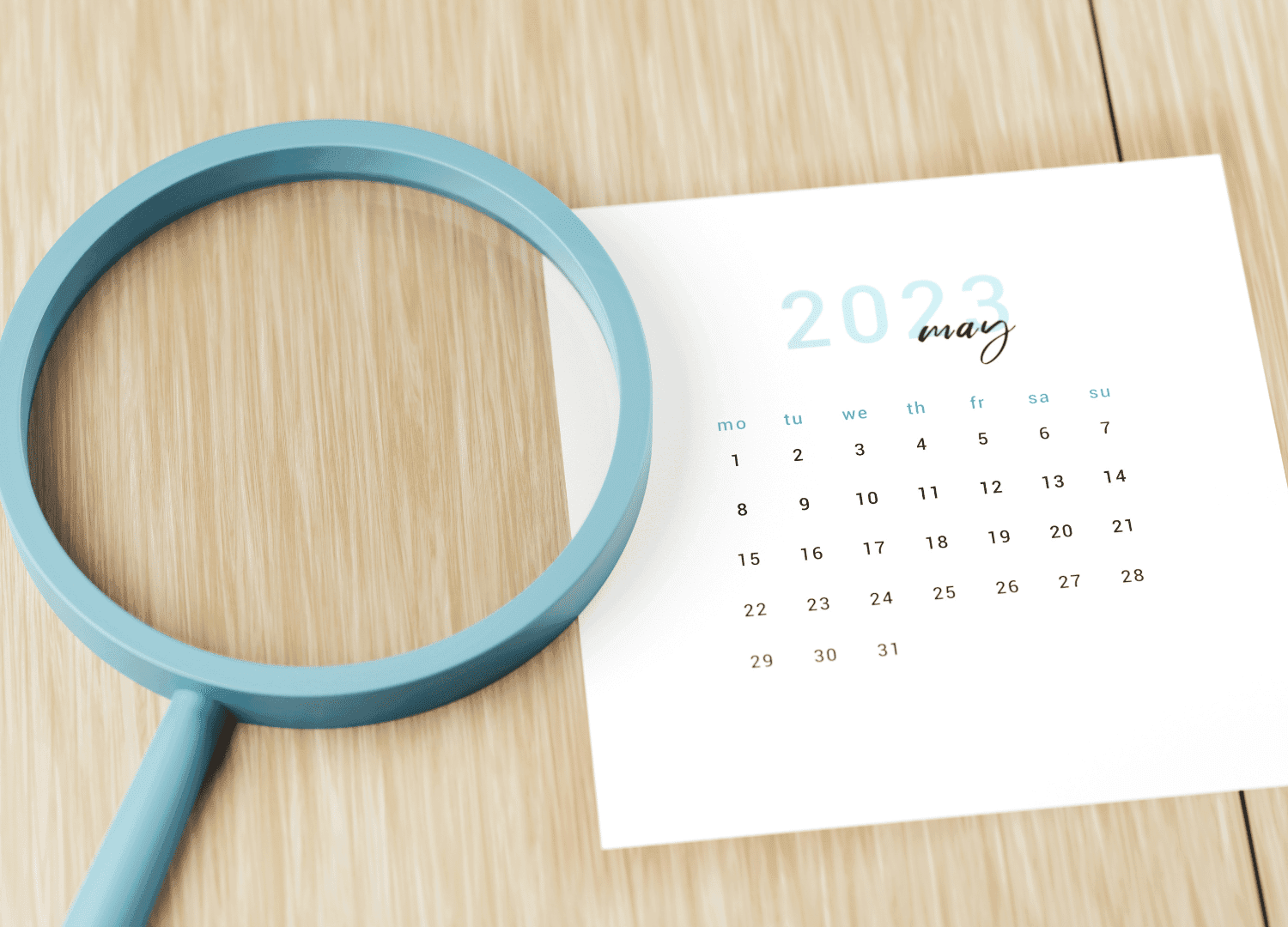 A blue magnifying glass rests beside a small calendar page showing May 2023, arranged on a light wooden surface. The calendar presents dates in a grid format, enhancing organization and clarity.