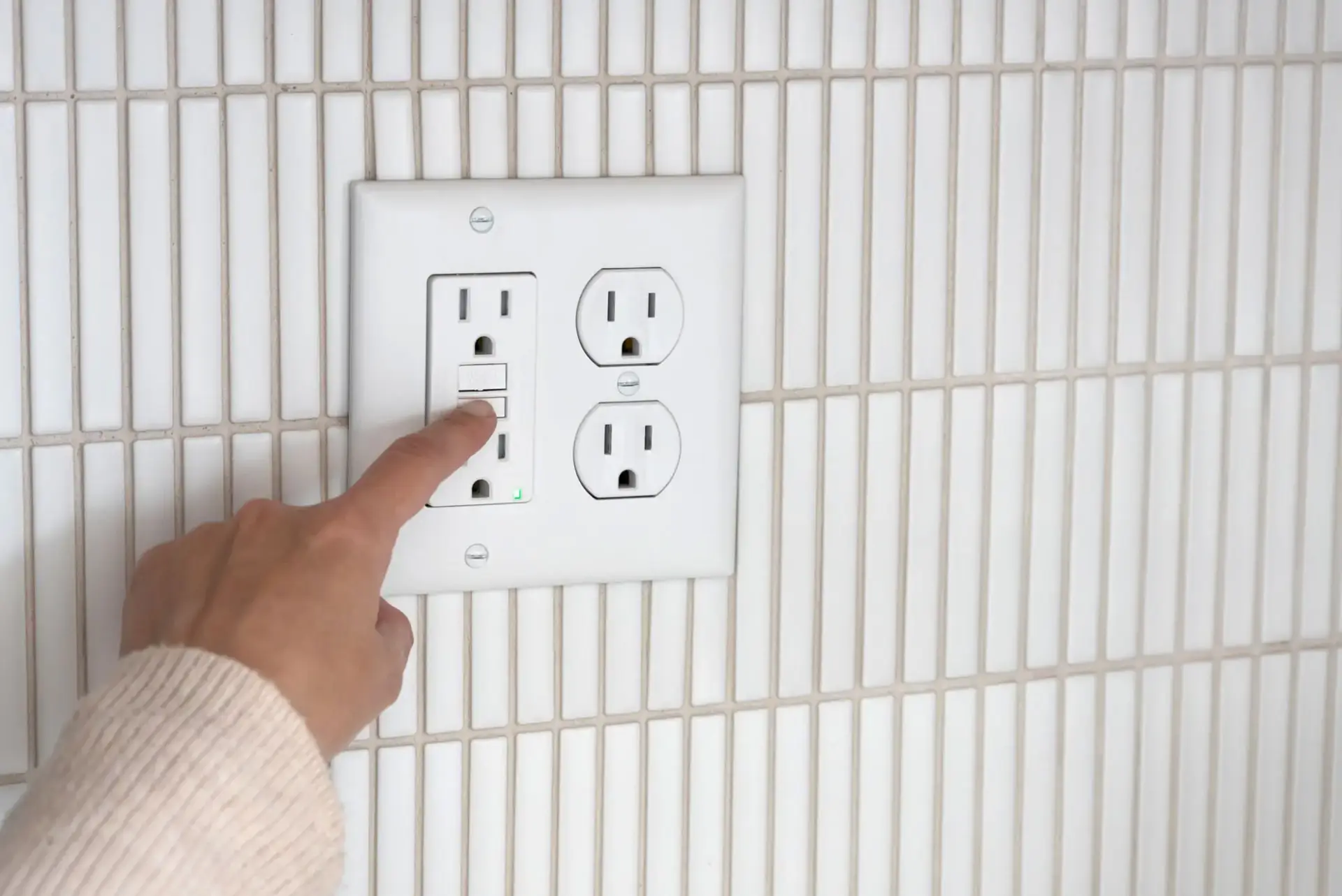 A hand in a beige sweater presses the test button on a white GFCI outlet mounted on a tiled wall, demonstrating how GFCI outlets work to enhance electrical safety.