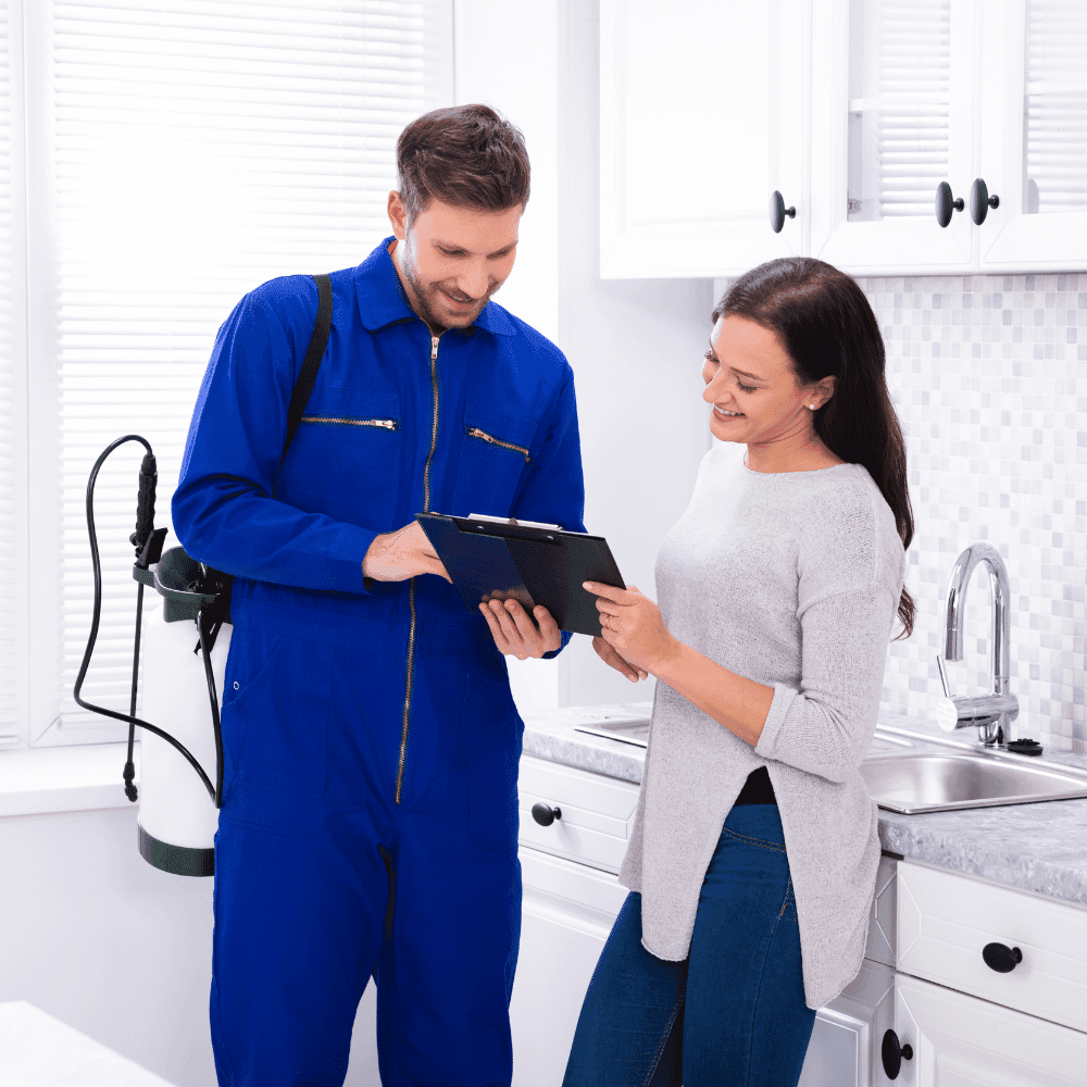 A pest control worker in a blue jumpsuit shows a digital tablet to a smiling woman in her kitchen, standing next to the sink and white cabinets—ideal for those searching for Pest Controllers Near Me.
