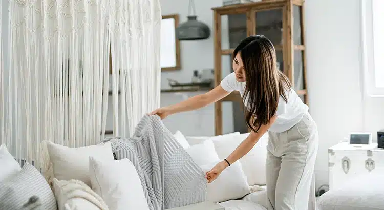 A woman in light clothing arranges pillows on a cream-colored couch in a bright, modern living room decorated with a macrame wall hanging and wooden furniture, creating an inviting atmosphere.