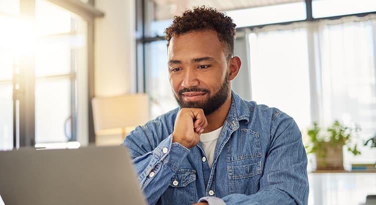 A man in a denim shirt sits at a desk, thoughtfully looking at his laptop in a bright, modern room filled with natural light and lush plants.