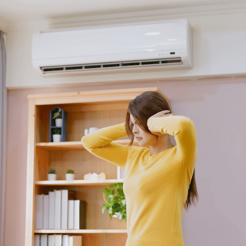 A woman in a yellow sweater stands indoors, holding her head with both hands and appearing stressed. Behind her, a bookshelf holds plants and books, while an air conditioner needing ac repair is mounted on the wall.