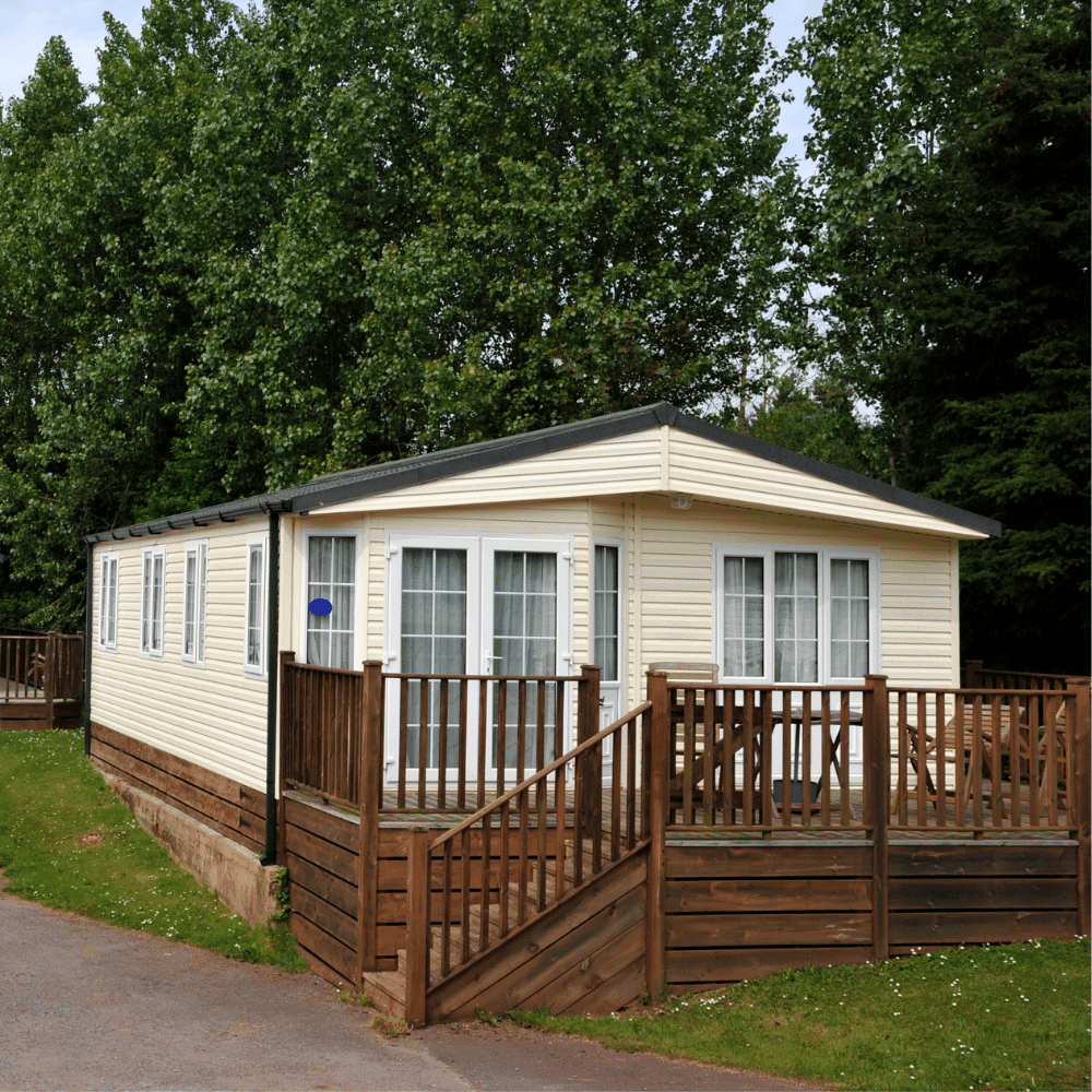 A cream-colored mobile home with large windows, visible ac units in the mobile home, and a wooden deck, surrounded by tall green trees on a grassy plot with a paved path in front.