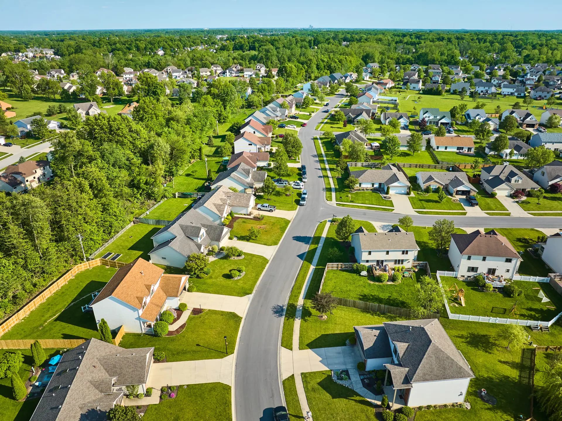 Aerial view of a suburban neighborhood with rows of houses, green lawns, trees, and winding streets under a clear blue sky on a sunny day, ideal for assessing properties during a Houston home inspection.
