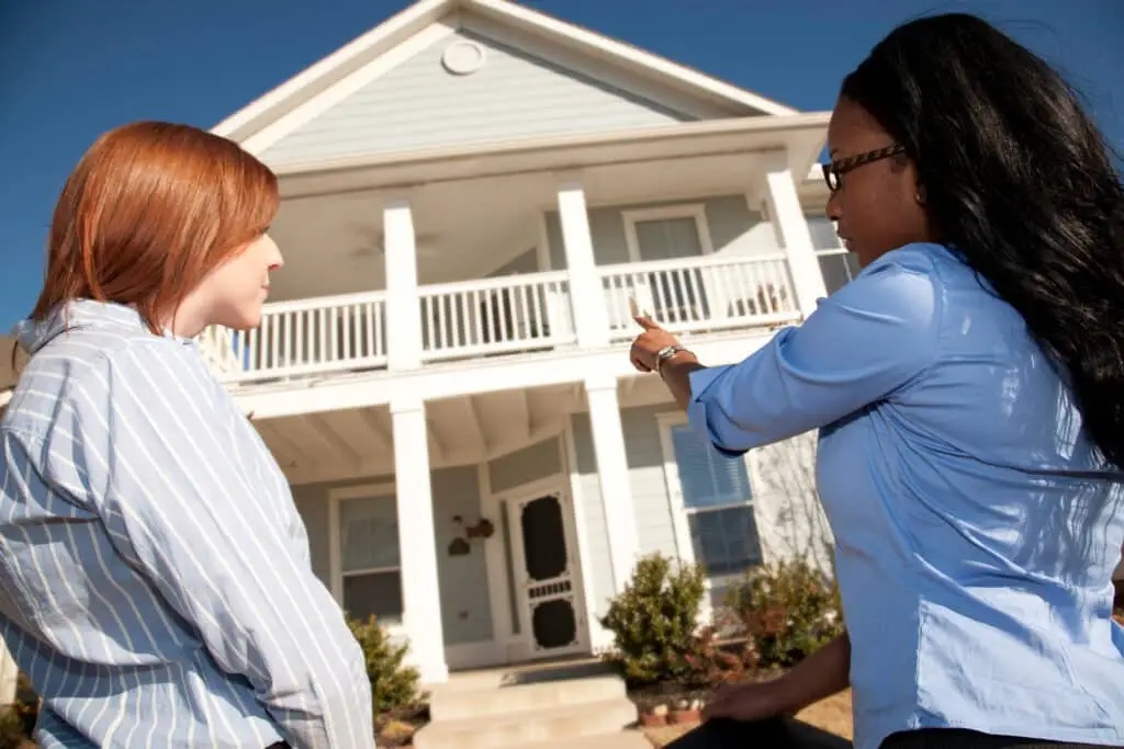 Two women stand outside a two-story house, one pointing towards the home—possibly discussing a first time homebuyer package on a sunny day. Shrubs and a pathway lead to the front door.