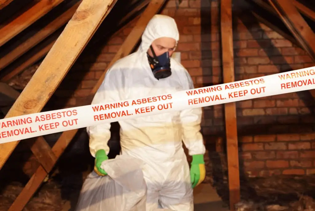 A person in protective gear and a respirator works in an attic behind tape labeled Warning Asbestos Removal. Keep Out, with exposed wooden beams and brick walls visible—similar precautions are taken during an asbestos inspection or radon assessment.