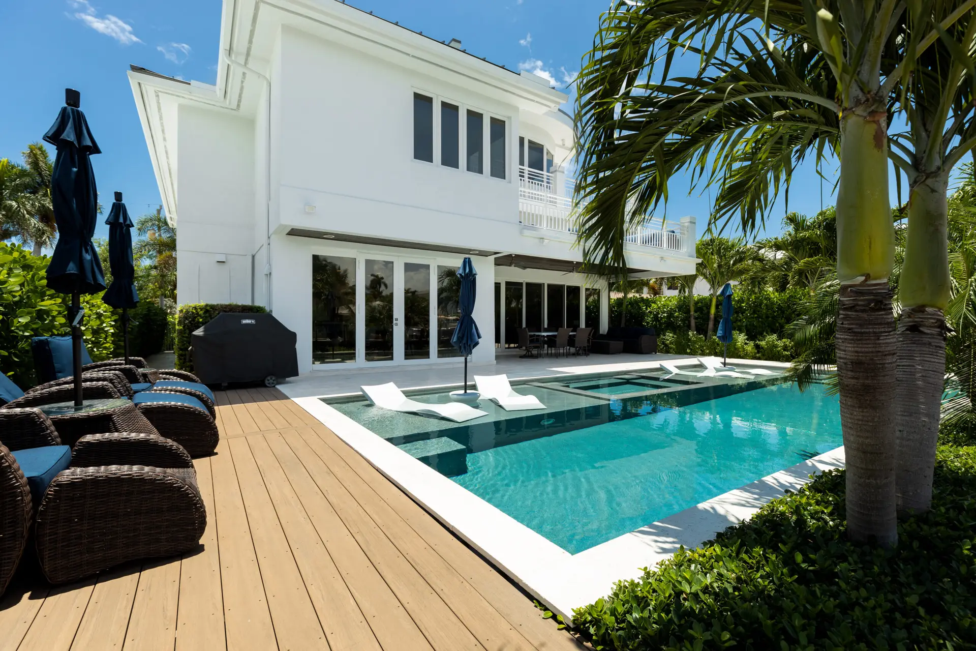 Modern white two-story house with large windows, a wooden deck, and a swimming pool featuring in-water lounge chairs—expertly reviewed by a Houston home inspector—surrounded by palm trees and outdoor furniture under a sunny sky.