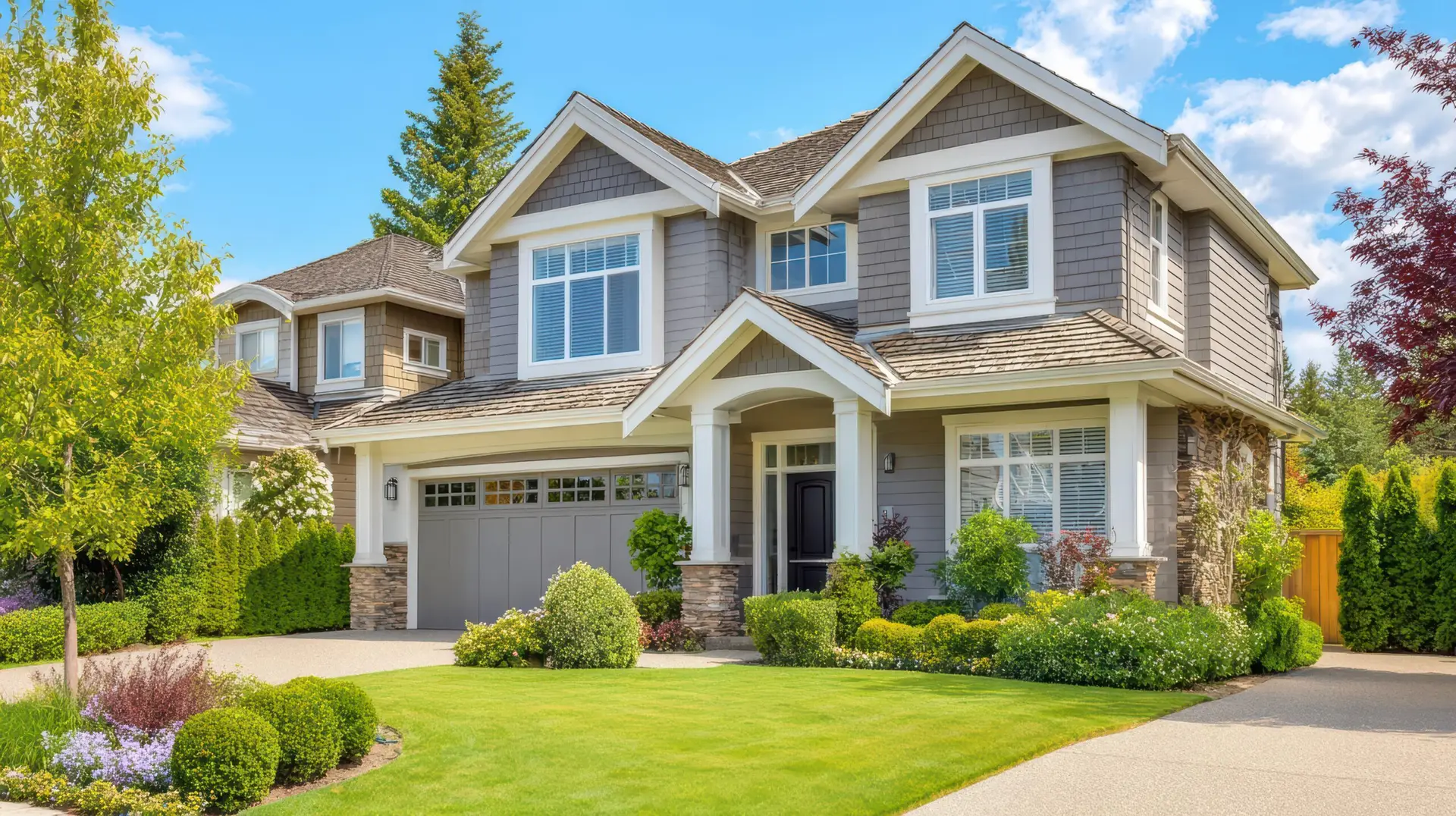 Two-story suburban house with gray siding, white trim, and stone accents. Neatly manicured lawn, shrubs, and colorful flowers surround the entrance—perfect for a Houston home inspection on a sunny day.