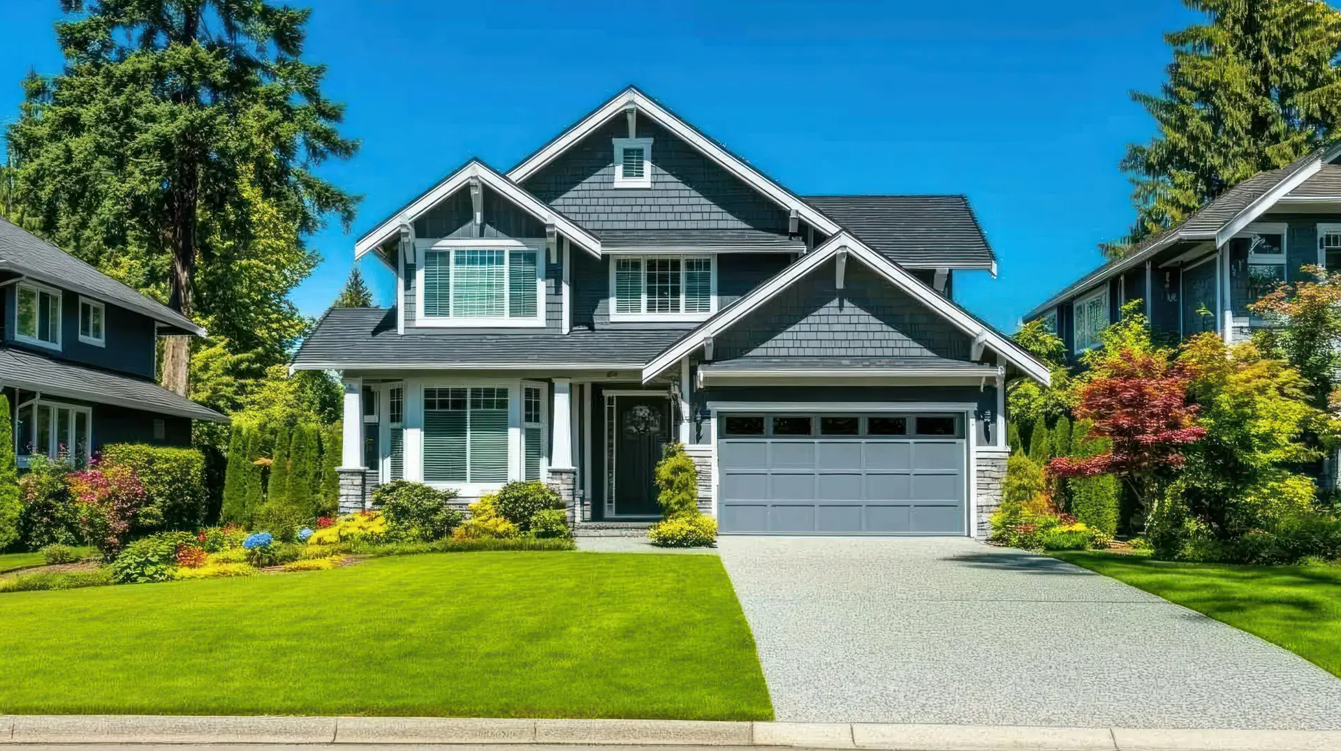 A modern two-story house with gray siding, white trim, and a double garage sits among lush green lawns, colorful shrubs, and tall trees under a bright blue sky—perfect for a Texas Real Estate home inspection.