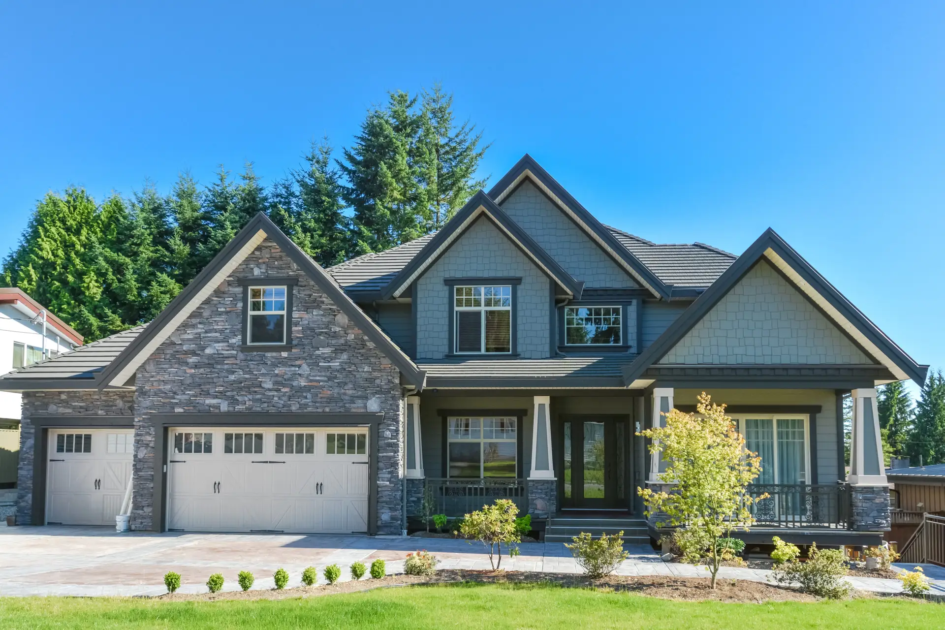 Modern two-story house with stone and gray siding exterior, three-car garage, large windows, and a covered front porch, surrounded by a well-kept lawn and trees under a clear blue sky—ideal for Texas Real Estate or your next home inspection.