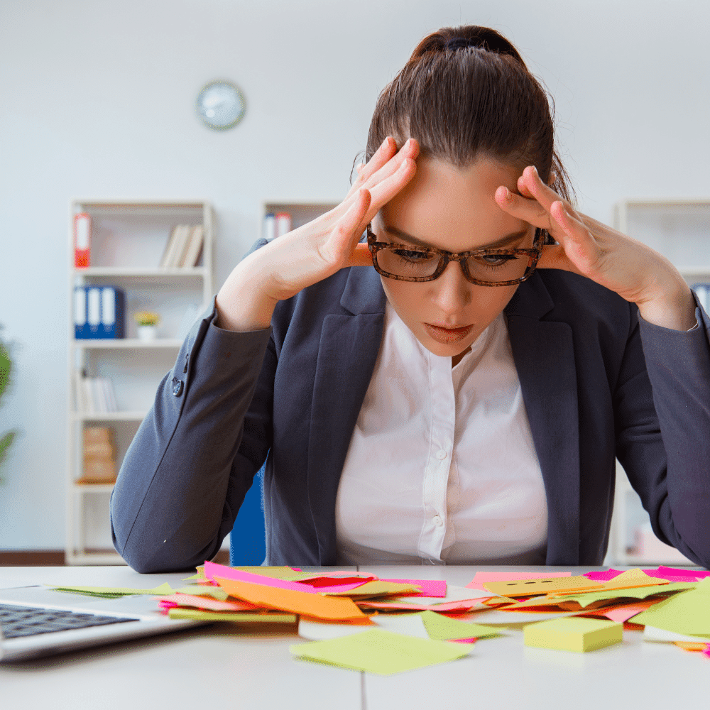 A woman in business attire sits at a desk covered with colorful sticky notes, holding her head in her hands and appearing stressed as she tries to schedule a home inspection in her busy office.