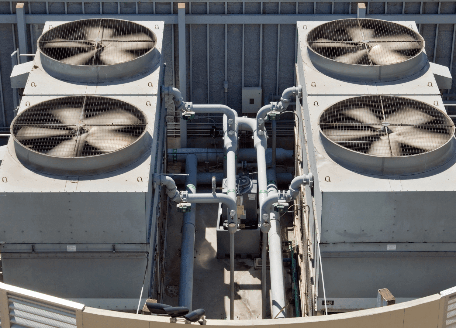 Four large industrial air conditioning units with spinning fans, metal piping, and grates are situated on a rooftop where technicians are checking AC pressure.