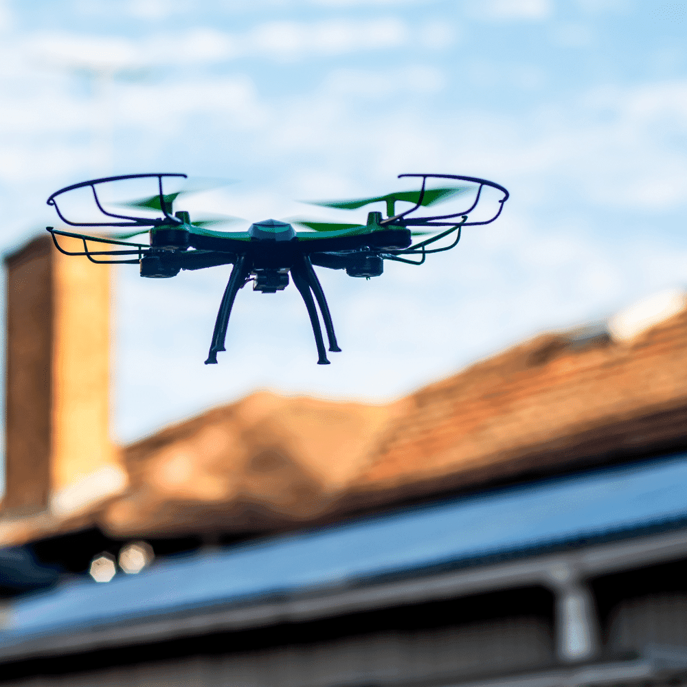A small black and green drone, used for drone inspection services, flies outdoors near rooftops under a blue sky with scattered clouds, with a brick chimney visible nearby.