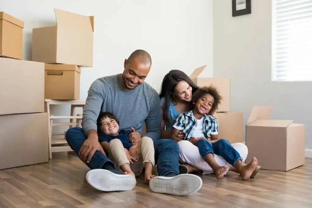 A happy family of four sits on the floor of their new home after a Texas home inspection, surrounded by moving boxes, smiling and laughing together.