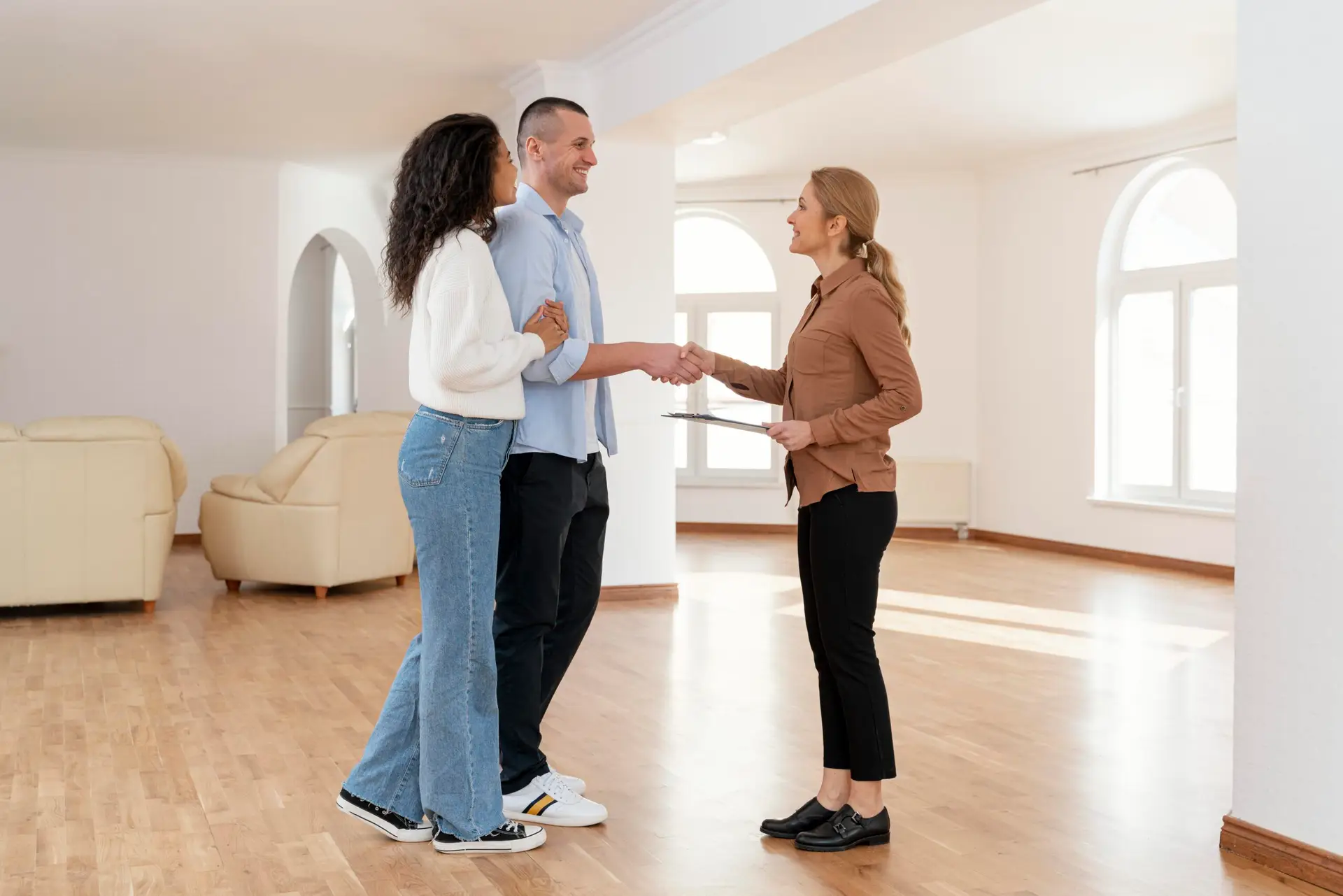 A couple stands arm in arm, smiling and shaking hands with a real estate agent inside a spacious living room with large windows and wooden floors, celebrating the value of their new real estate investment.