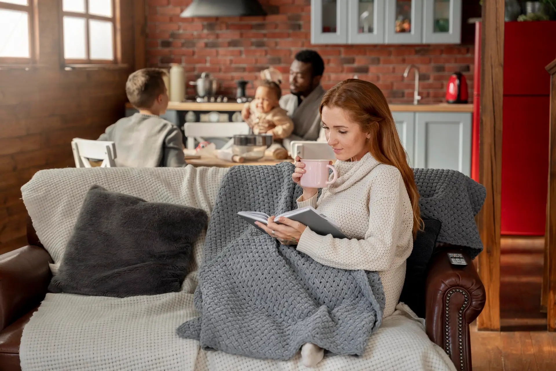 A woman sits on a cozy couch with a blanket, reading a book and drinking from a mug. The warm, homey setting helps keep heat in as a man and two children enjoy time together at the kitchen table.