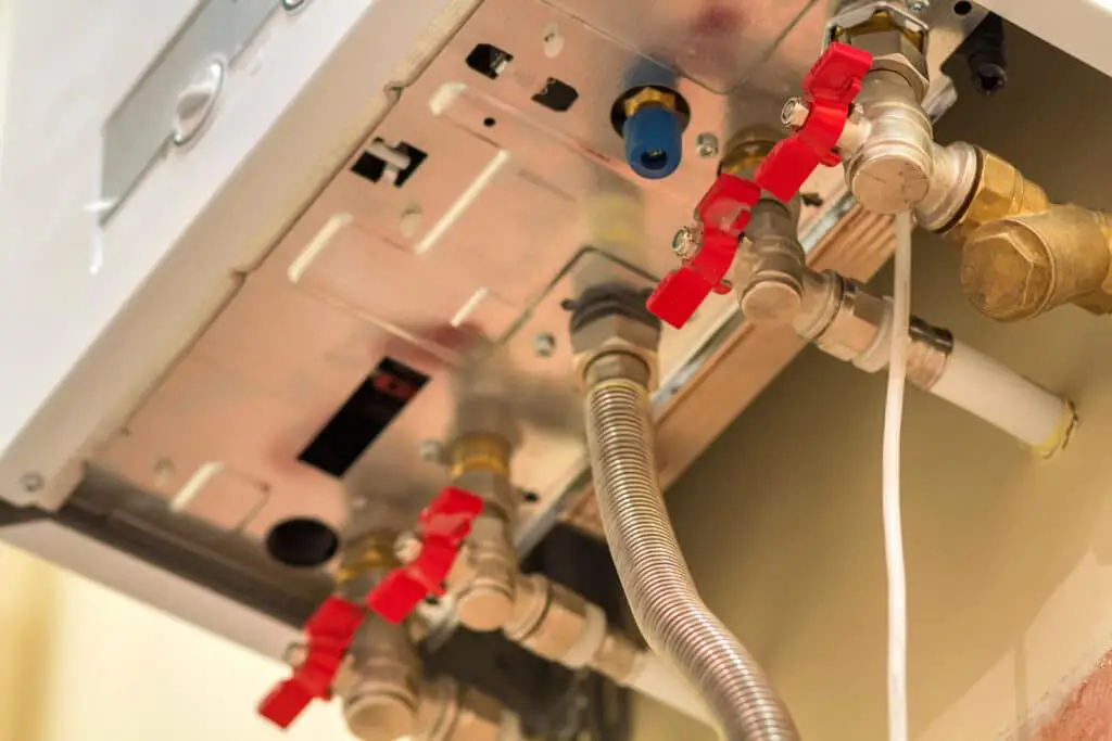 Close-up view of the underside of a gas boiler showing multiple pipes, valves with red handles, and flexible metal connections. This setup is useful when learning how to adjust temperature on a hot water heater.
