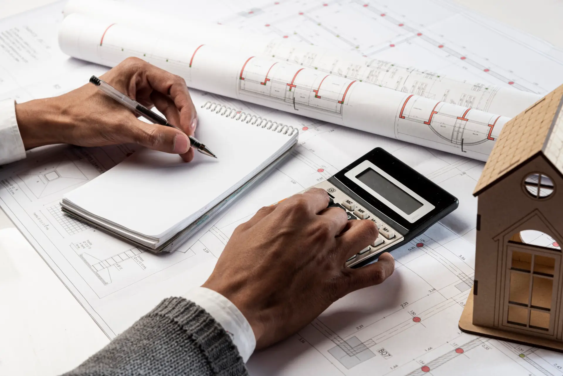 A person writes in a notebook with one hand while using a calculator with the other, surrounded by architectural blueprints and a small wooden house model, estimating the cost of home inspection 2025.