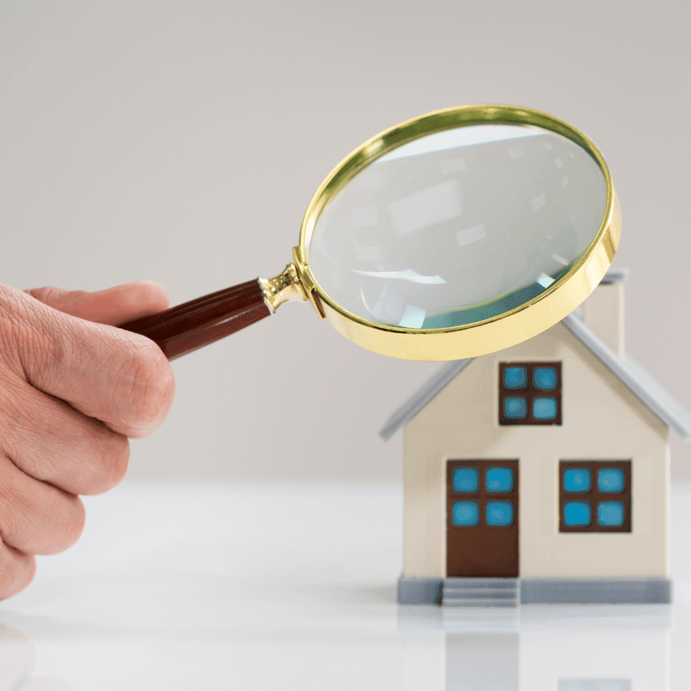 A hand holding a magnifying glass closely examines a small model of a house with blue windows and a brown door, symbolizing home inspection or real estate evaluation using tools like a zip level.