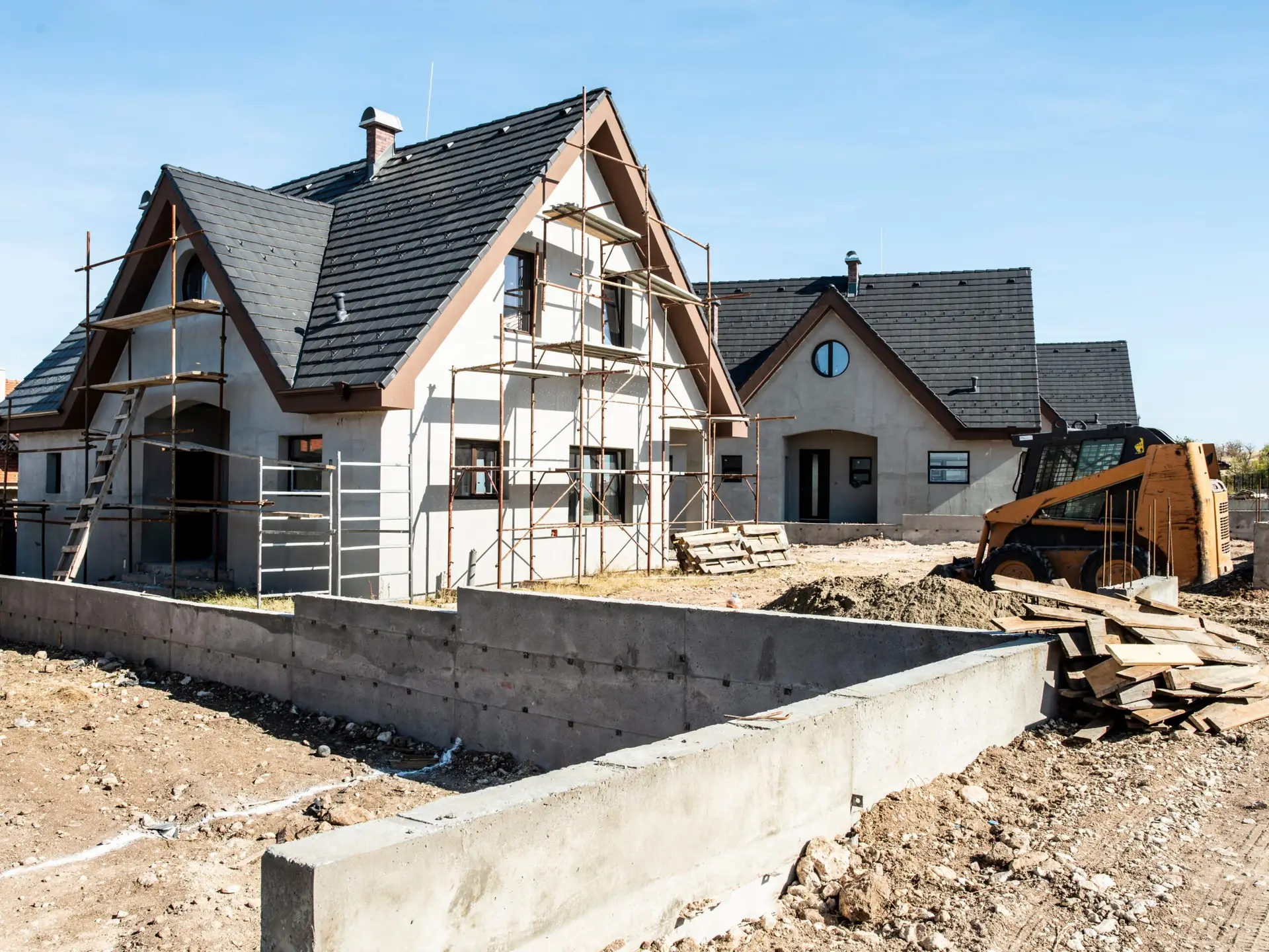 Two houses under construction with scaffolding and unfinished exteriors are surrounded by dirt and construction materials. A yellow loader is parked nearby, and a concrete foundation is visible—ideal conditions for a Texas home inspection.