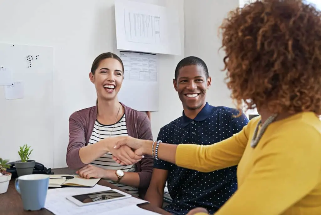 Three people sit at a desk in an office during a post-inspection negotiation, two facing the camera and smiling as the third, with curly hair, shakes hands with one of them. Papers, a tablet, and coffee cups are on the table.