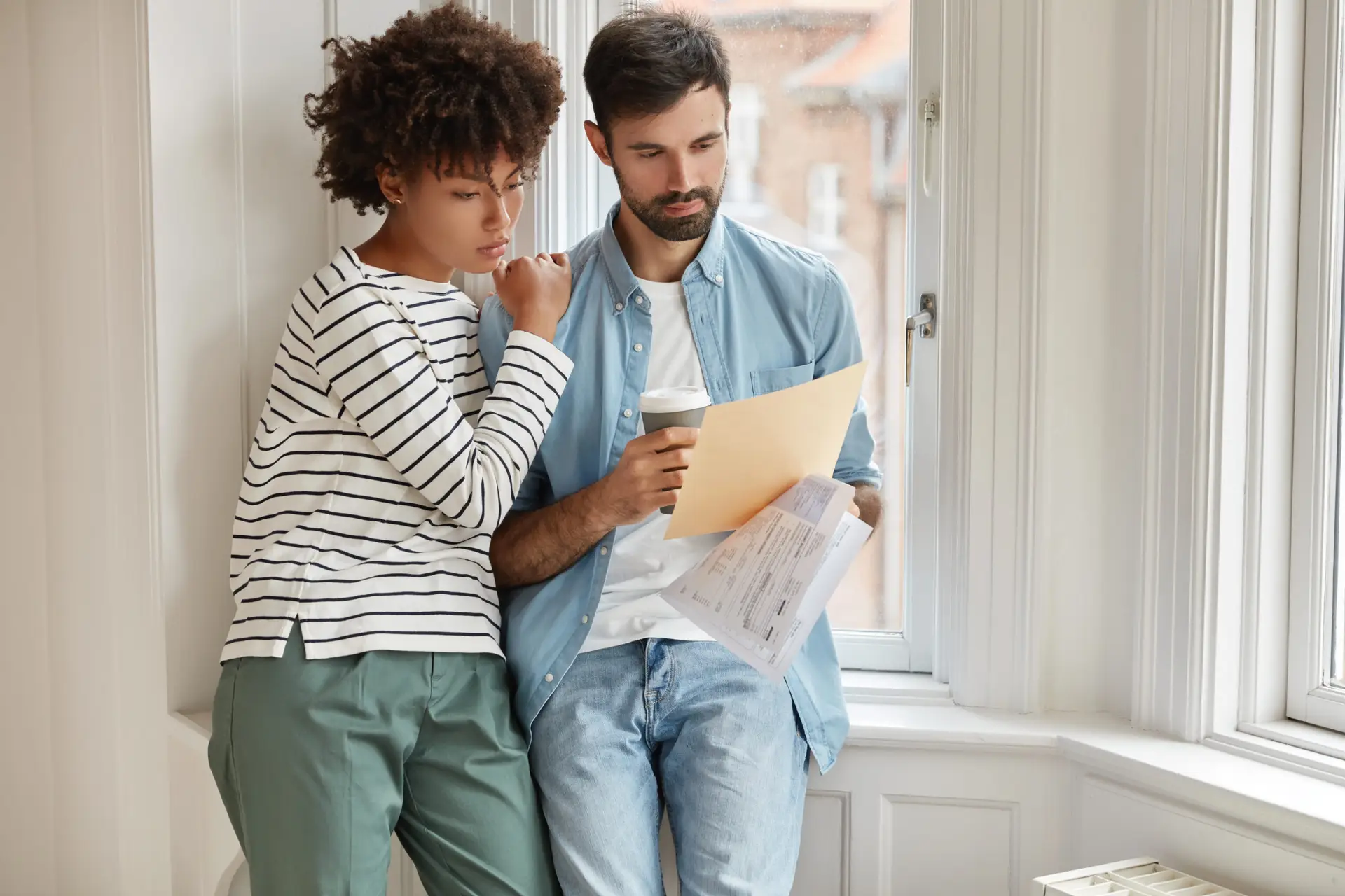 A woman and a man stand by a window, reviewing documents together. The woman appears thoughtful, while the man holds papers and a coffee cup. Both seem focused and concerned, possibly discussing findings from a home inspection company.