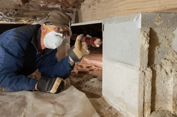 A person wearing gloves, a hat, and a protective mask inspects a building's foundation in a crawl space, shining a flashlight on the structure during a home inspection service by Super Home Services.