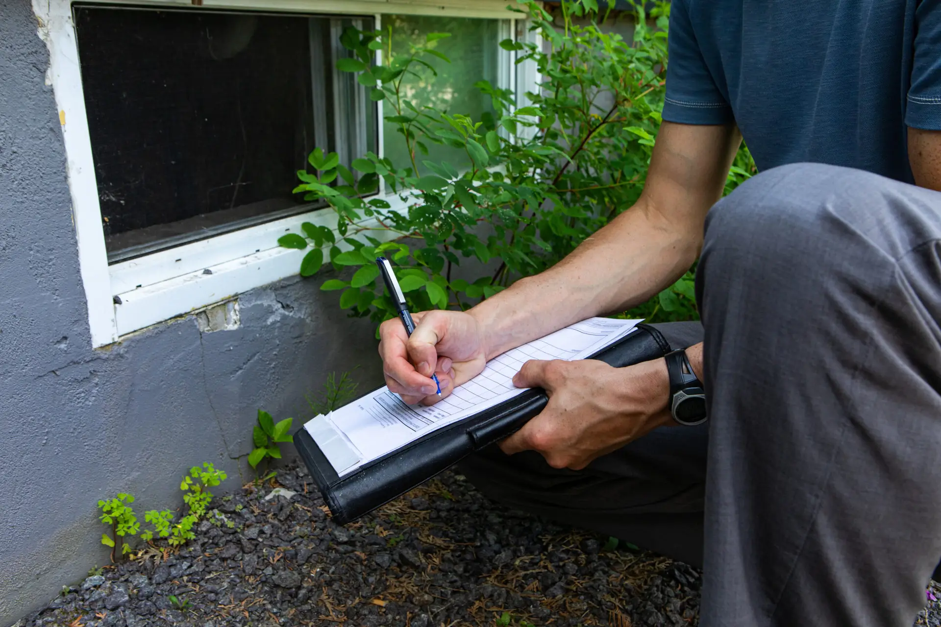 A Houston home inspector kneels outside next to a building, writing on a clipboard near a basement window surrounded by plants and gravel. Only their torso, arms, and legs are visible.