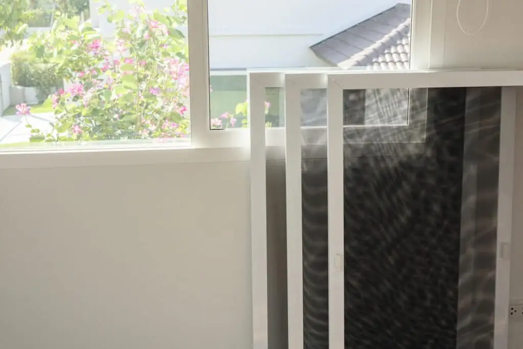 Three window screens rest against a white wall below an open window, with sunlight casting patterns and a view of greenery and flowers—an inviting scene often noted during home inspections.