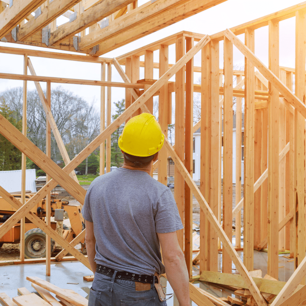 A construction worker wearing a yellow hard hat and gray shirt stands at the frame of new construction homes, observing the building progress on a sunny day.