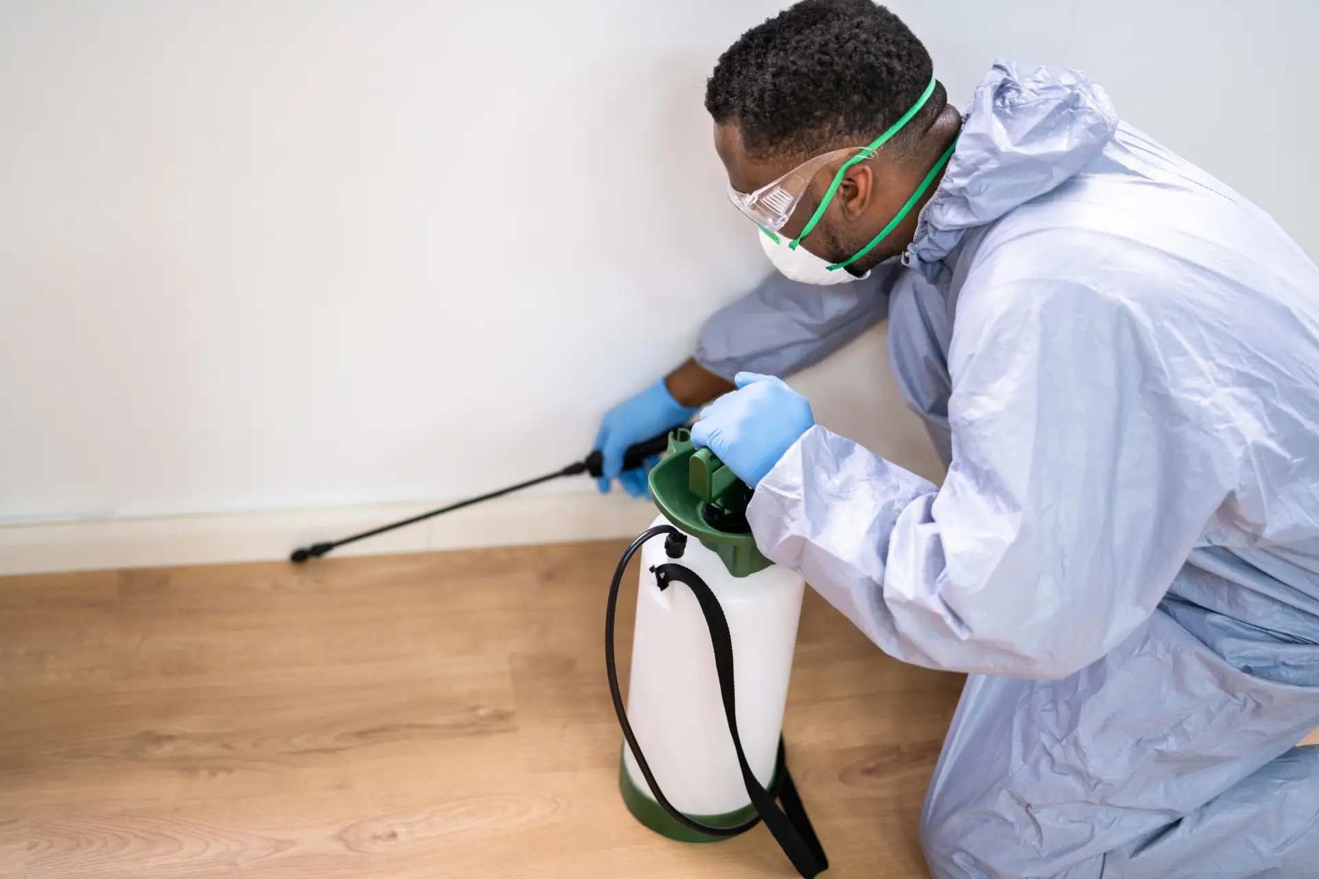 A person in protective gear and gloves uses a hand-held sprayer to apply pesticide along the base of a white wall on a wooden floor—perfect for those seeking Cyber Monday Home Utility deals.