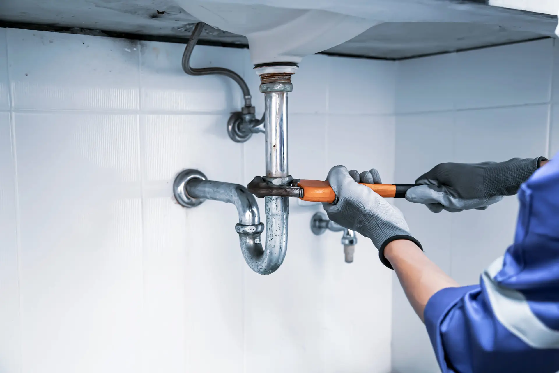 A plumber wearing gloves uses a pipe wrench to fix or tighten the curved drain pipe under a sink, working on a blocked kitchen sink against a white tiled wall.