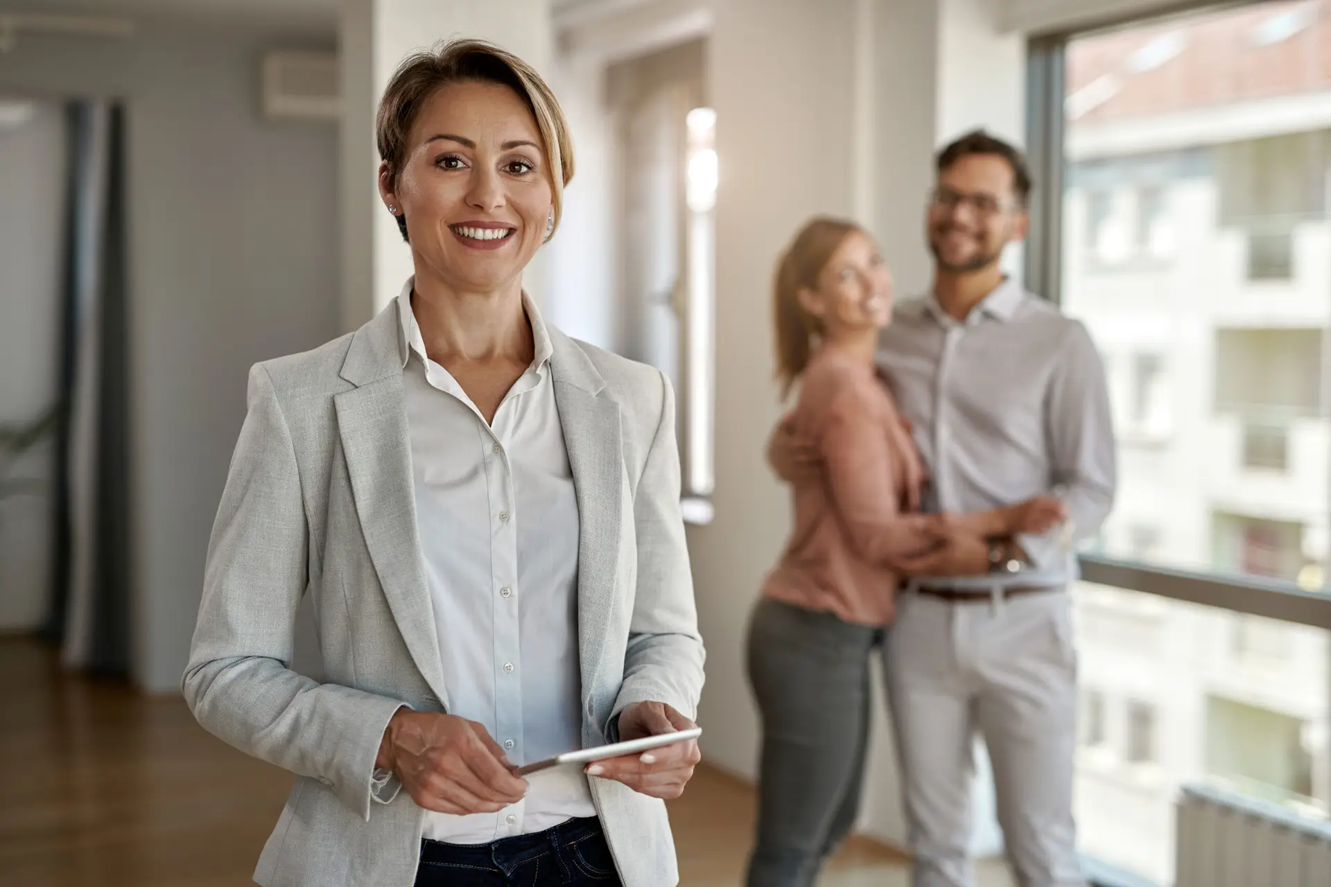 A smiling real estate agent holding a tablet, enhanced by real estate technology, stands in the foreground as a happy couple hugs inside a bright, modern apartment.