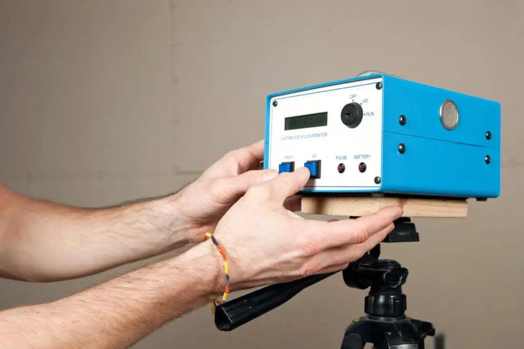 A person adjusts the knob on a blue electronic device with a display and several buttons, mounted on a tripod—possibly used for integrated pest management. The person is wearing a colorful thread bracelet on their wrist.
