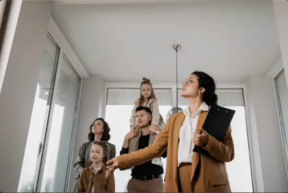 A real estate agent in a brown suit shows a modern, bright home to a family of four, discussing real estate predictions 2024 as the family explores and the agent highlights key features.