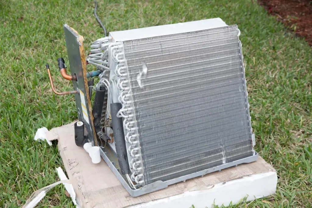 A central air conditioning evaporator coil unit placed on cardboard on grass, showing coiled tubing and metal fins, with some frost and copper pipes visible—an issue like a burning smell when I turn the heat on may occur in such systems.