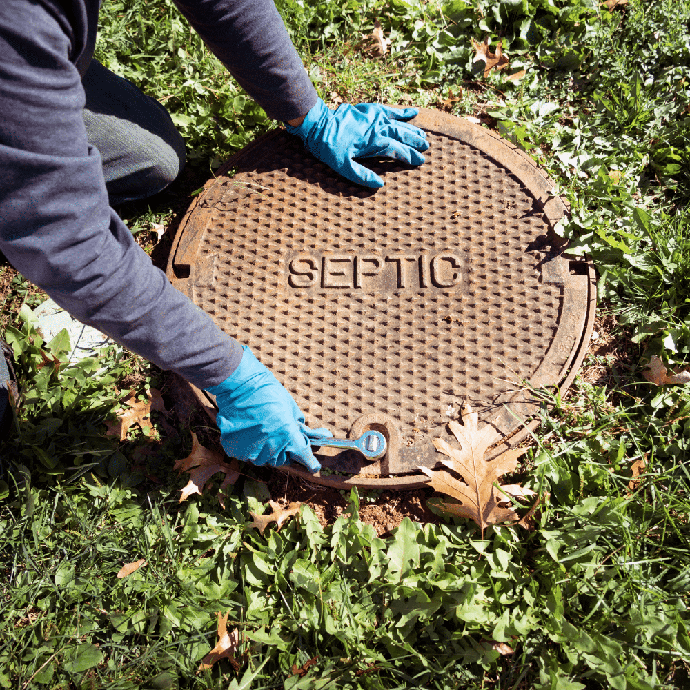 A person in blue gloves uses a wrench to open a round septic tank cover labeled SEPTIC, surrounded by grass and fallen leaves, performing septic inspections.