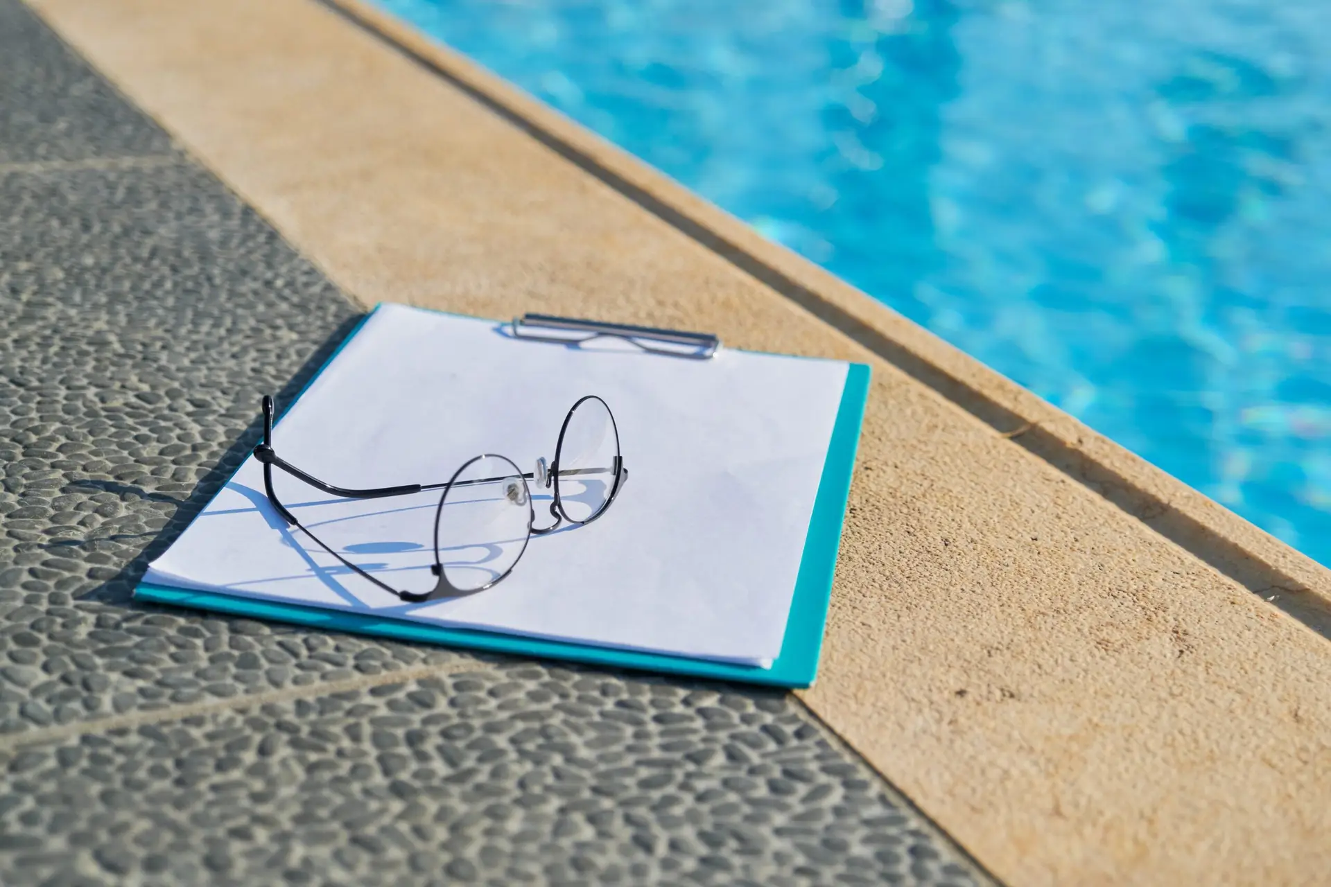 A clipboard with blank paper and a pair of eyeglasses rests on the edge of a swimming pool, hinting at Home Inspections in Frisco—where attention to detail is key for any home inspection company.