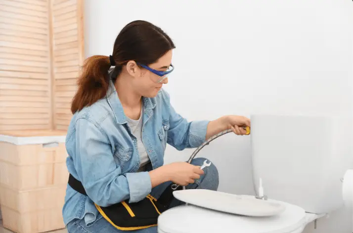 A woman wearing safety glasses and a tool belt uses a wrench to repair a toilet tank with the lid removed, demonstrating how to fix a wobbly toilet by tightening the plumbing connection.